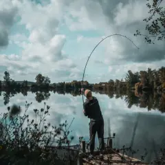 Wędkarz na pomoście walczy z rybą na spokojnej rzece. W tle drzewa odbijają się w tafli wody.