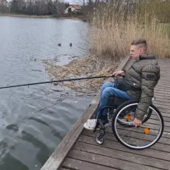 Młody mężczyzna na wózku inwalidzkim łowi ryby na drewnianym pomoście nad jeziorem w Iławie.