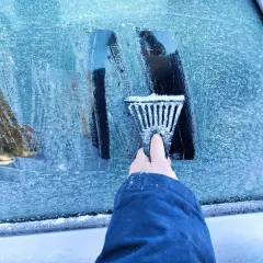 Ręka z **przeręblem** usuwa lód z zamarzniętej szyby samochodu.