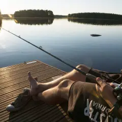 Słońce zachodzi nad spokojnym jeziorem. Wędkarz siedzi na pomoście, trzymając wędkę, gotowy na złapanie ryby.