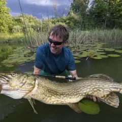 Szczęśliwy wędkarz trzyma ogromnego szczupaka, prawdopodobnie największego szczupaka na świecie, w wodzie wśród lilijek.