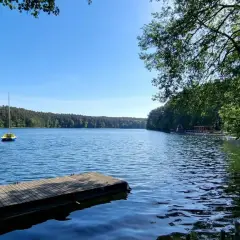 Drewniane pomosty nad spokojnym jeziorem, z małą żółtą łódką na wodzie i lasem w tle. Słońce odbija się od powierzchni, tworząc wrażenie, jakby jezioro było "spalone" blaskiem.