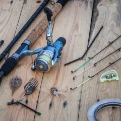 Wędki feeder ranking: zestaw sprzętu wędkarskiego z kołowrotkiem, ciężarkami, sygnalizatorami brań i żyłką na drewnianym tle.