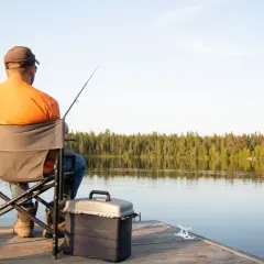 Mężczyzna z wędką siedzi na pomoście. Obok niego stoi pudełko na sprzęt, a w tle rozciąga się las nad jeziorem. Posiada ważną legitymację wędkarską.
