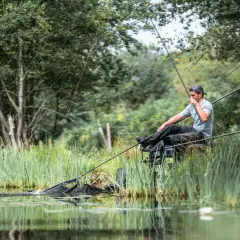 Wędkarz wyciąga rybę z wody, używając podbieraka. W tle bujna zieleń i woda, a na pierwszym planie widać cienką, pomarańczową linę.