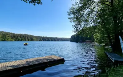 Drewniane pomosty nad spokojnym jeziorem, z małą żółtą łódką na wodzie i lasem w tle. Słońce odbija się od powierzchni, tworząc wrażenie, jakby jezioro było "spalone" blaskiem.