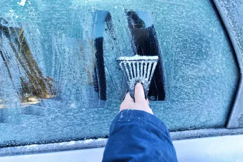 Ręka z **przeręblem** usuwa lód z zamarzniętej szyby samochodu.