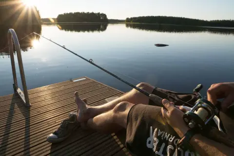 Słońce zachodzi nad spokojnym jeziorem. Wędkarz siedzi na pomoście, trzymając wędkę, gotowy na złapanie ryby.