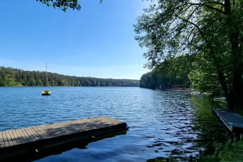 Drewniane pomosty nad spokojnym jeziorem, z małą żółtą łódką na wodzie i lasem w tle. Słońce odbija się od powierzchni, tworząc wrażenie, jakby jezioro było "spalone" blaskiem.