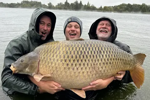 Trzech wędkarzy z dumą prezentuje gigantycznego karpia, prawdopodobnie nowy rekord. Ryba jest ogromna, a jej łuski lśnią w wodzie.