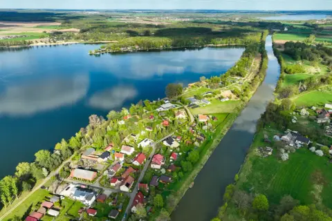 Malownicze pławniowice jezioro otoczone zielenią i domkami letniskowymi. Woda odbija chmury, a kanał przecina krajobraz.