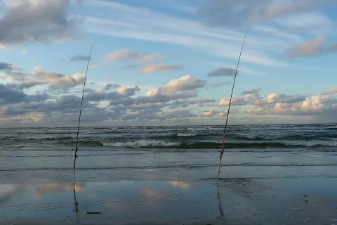 Dwie wędki stoją na plaży, czekając na moment, kiedy bierze szczupak. Morze faluje pod błękitnym niebem z chmurami.