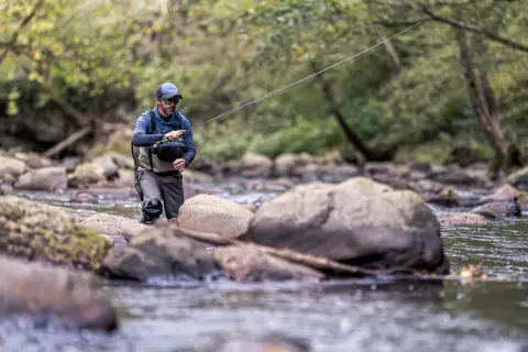 Wędkarz w kamizelce łowi w górskim potoku, licząc na złapanie pstrąga strumieniowego.