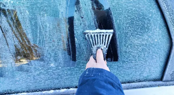 Ręka z **przeręblem** usuwa lód z zamarzniętej szyby samochodu.