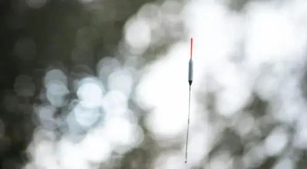 Szary spławik 5g z czerwoną końcówką, zawieszony na żyłce, na tle rozmytych, jasnych kółek.