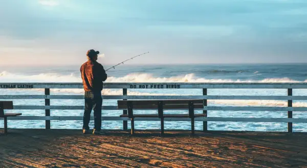 Mężczyzna z wędką na molo, cieszący się spokojnym dniem wędkarza nad wzburzonym oceanem.