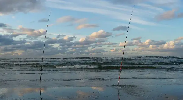 Dwie wędki stoją na plaży, czekając na moment, kiedy bierze szczupak. Morze faluje pod błękitnym niebem z chmurami.