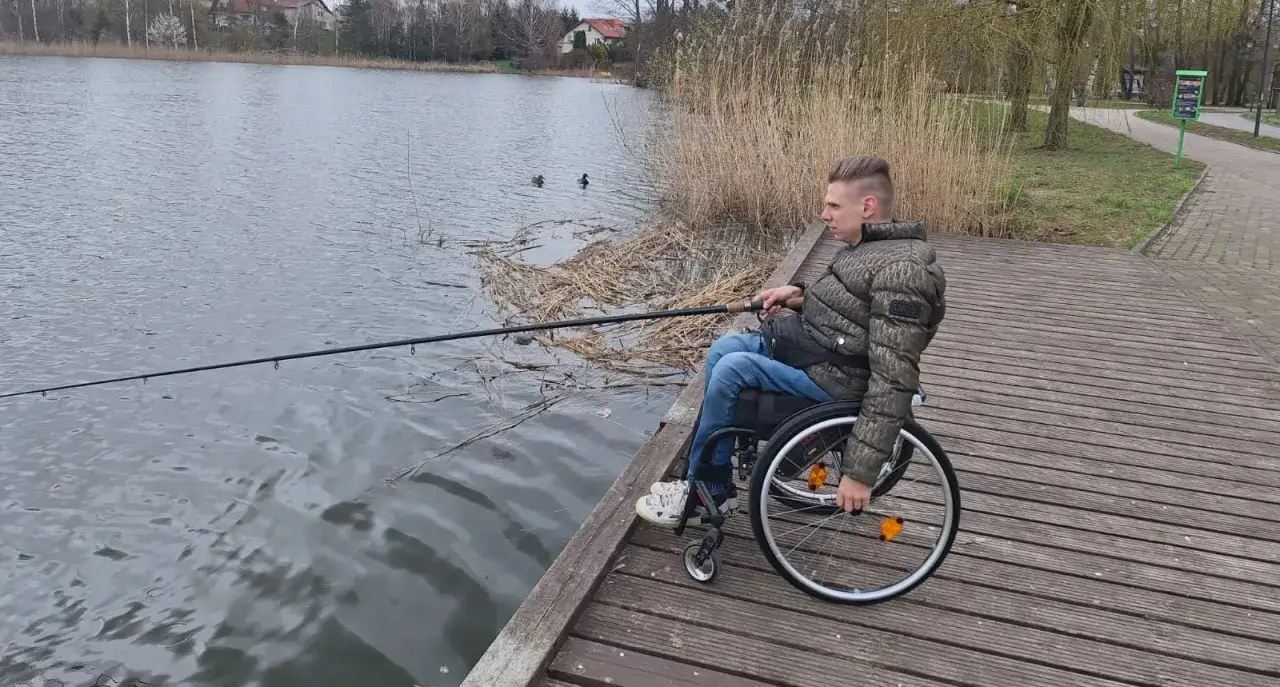 Młody mężczyzna na wózku inwalidzkim łowi ryby na drewnianym pomoście nad jeziorem w Iławie.