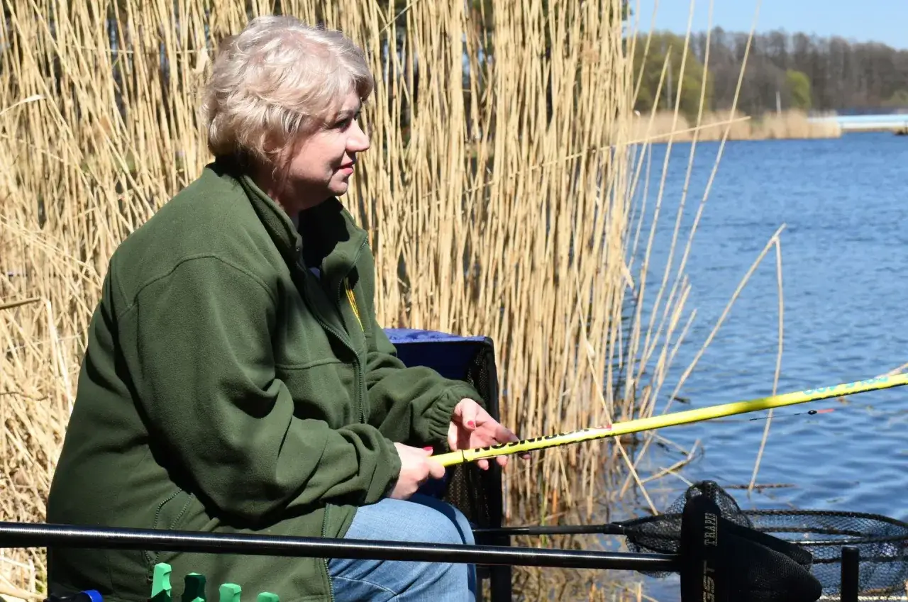 Kobieta łowi ryby nad jeziorem, ciesząc się spokojem. Wędka i podbierak wskazują, że jest to pasjonatka, która dba o polskie wody.