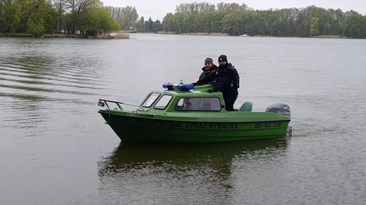 Dwóch funkcjonariuszy Państwowej Straży Rybackiej na zielonej łodzi patrolowej na jeziorze.