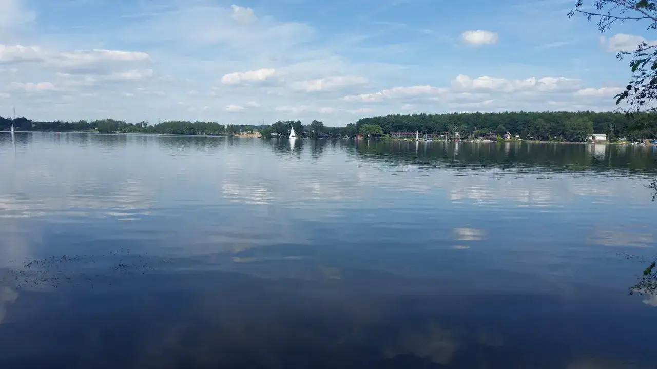 Spokojne jezioro Pławniowice z odbijającymi się chmurami i żaglówkami na horyzoncie.