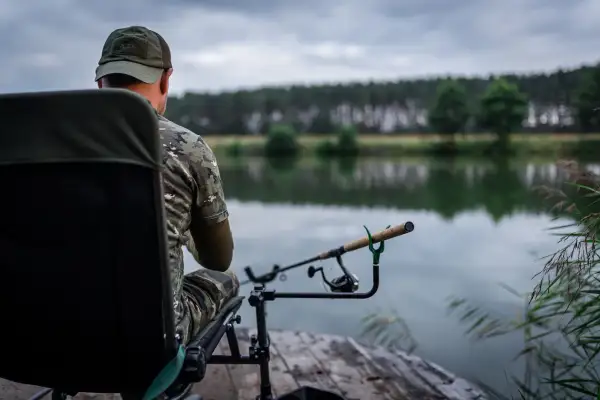 Wędkarz w kamuflażu czeka na branie, opierając swoje wędziska na stojaku nad spokojną wodą.