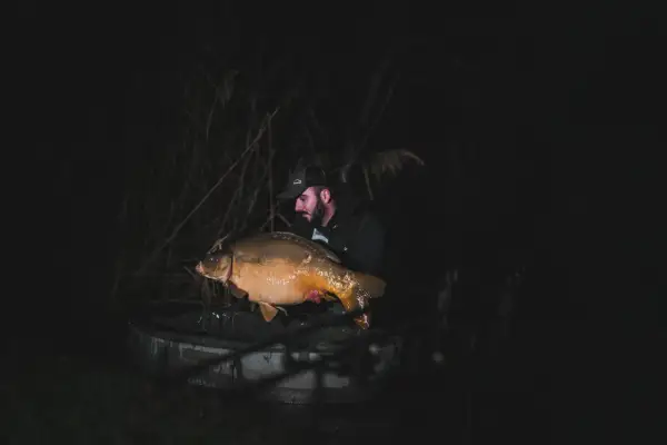 Wędkarz z dumą prezentuje ogromnego karpia, którego udało mu się złowić nocą. To niezapomniane chwile, kiedy biorą ryby.